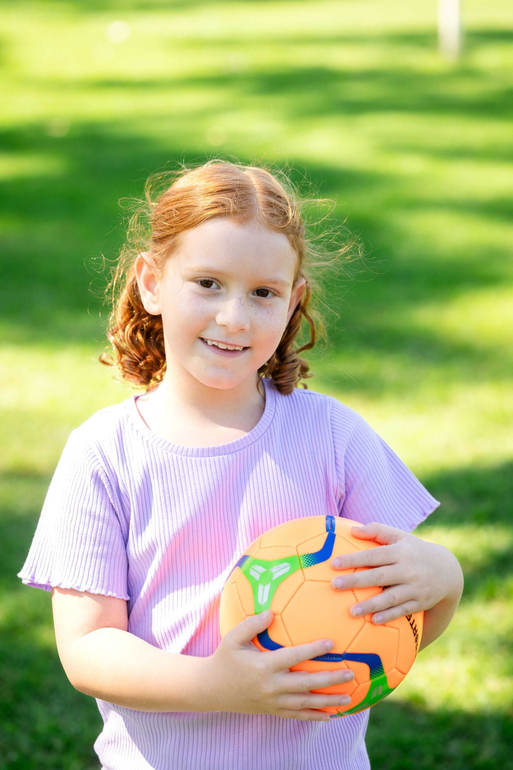 Small girl with red hair and pink shirt outside on grass smiling and holding orange ball