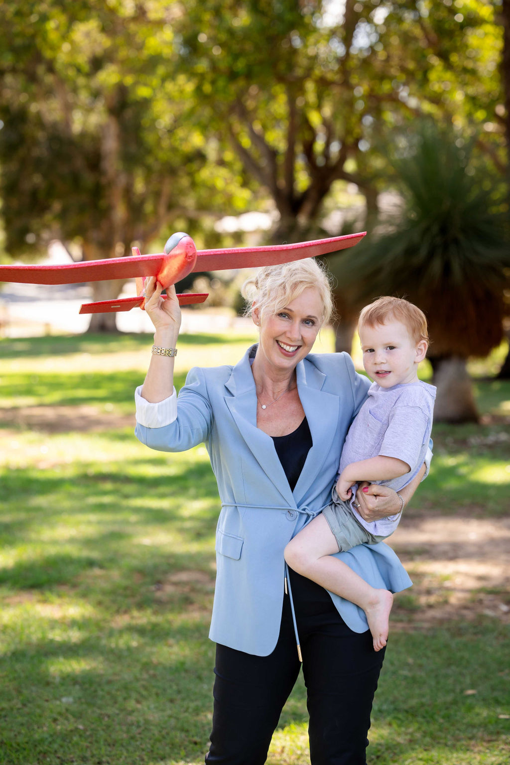 Jackie Davis with a toy aero plane and a little boy on her hip in a park